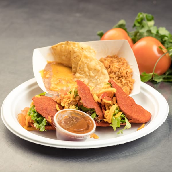 Two red tacos with lettuce, cheese, and meat, served with rice, beans, chips, and salsa.