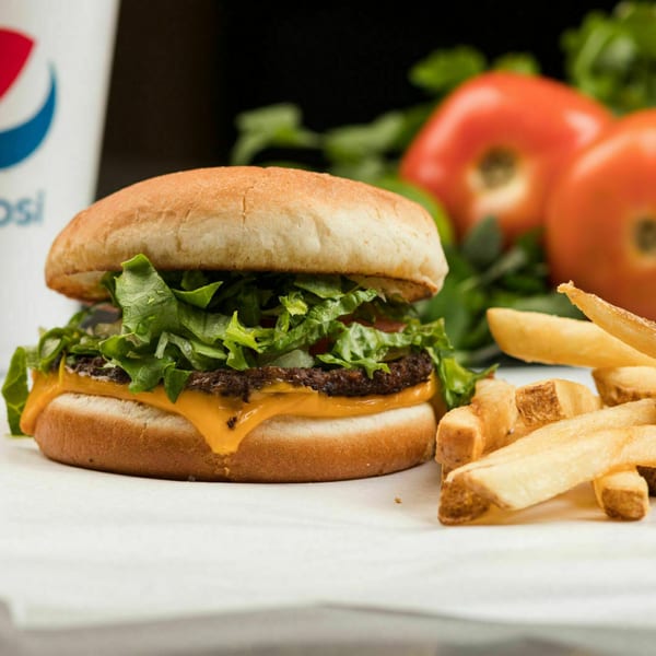 Cheeseburger with lettuce, fries, a Pepsi cup, and fresh tomatoes on a white surface.